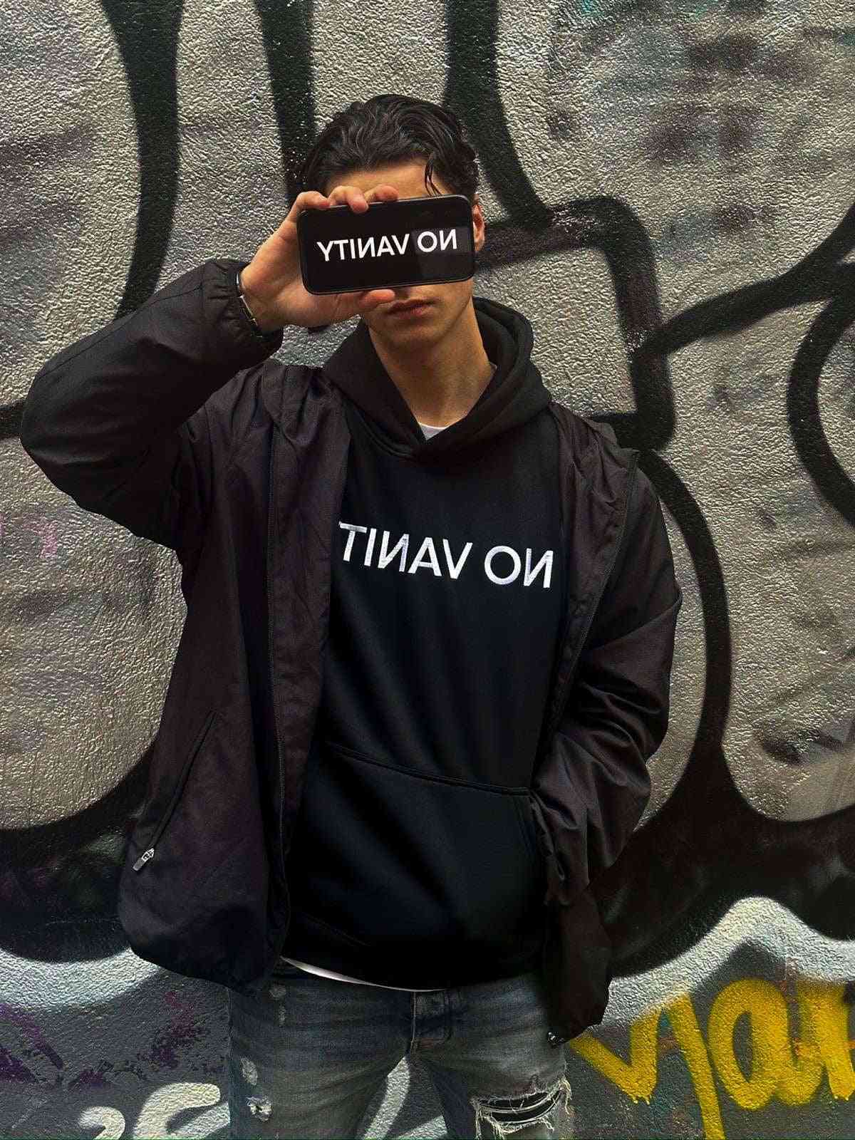 REVERSE NOVANITY HOODIE in black with mirrored 'NO VANITY' text on front, styled with a black jacket and jeans against graffiti wall.